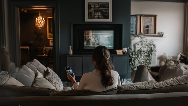 A woman with long dark hair sits from behind on a pale sofa in a dim living room, holding a lit phone, facing a wall-mounted television