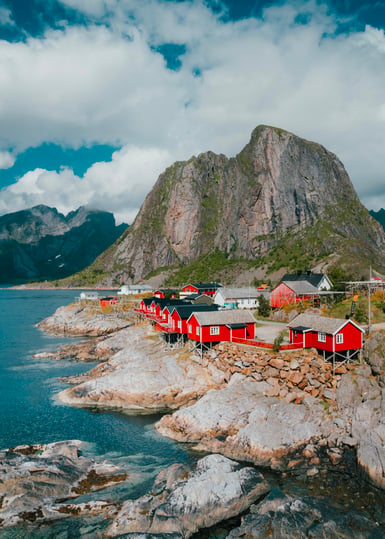 A cluster of red wooden cabins on a rocky shoreline beside blue water, with a steep grey mountain and cumulus clouds behind them