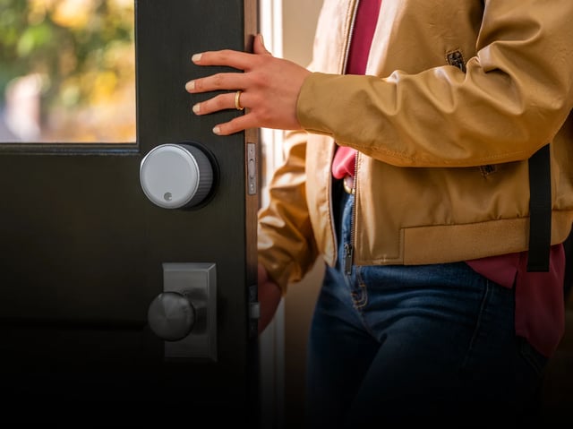 A person in a tan jacket and blue jeans pushes open a dark door fitted with a round white smart lock above a black euro cylinder knob