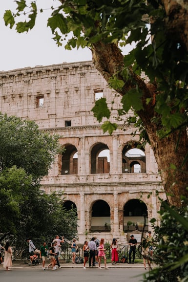 The tiered stone arches of the Colosseum in Rome rise behind a line of tourists on a pavement, framed at right by a plane tree trunk and overhanging leaves
