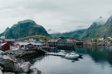 A small Lofoten fishing village of red and white wooden houses on stilts beside still harbour water, with two white boats moored and cloud-covered green mountains behind