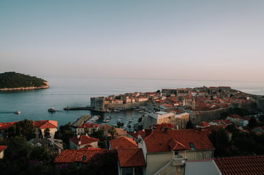Elevated view over Dubrovnik's walled old town on a headland by a calm sea, with red-tiled rooftops in the foreground, a small harbour of boats, and a wooded island offshore under a pale sky