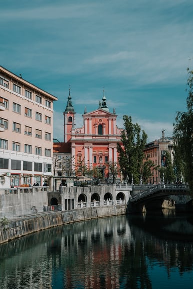 A pink baroque church with twin bell towers rises behind a stone-balustraded bridge and riverside buildings, reflected in the still green water of a canal lined with willows