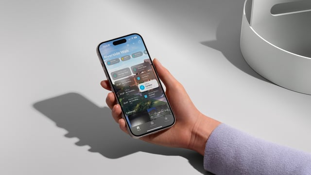 A hand in a lavender sleeve holds an iPhone displaying a home control app with tiles, casting a sharp shadow on a white surface beside a grey bowl