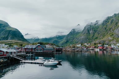 A Lofoten fishing village of red and white wooden houses on stilts around a still harbour, with small white boats moored at a jetty and cloud-covered green mountains behind