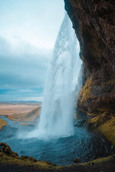 A tall waterfall drops from an overhanging mossy cliff into a shallow pool, with the flat, straw-coloured Icelandic lowland and a small farm building visible past the falling water