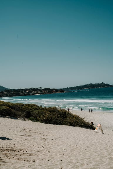 A wide white sand beach with dune scrub in the foreground, a few small figures walking near breaking waves, turquoise bay water, and a wooded headland with houses under clear blue sky