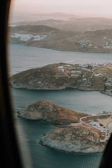 Aerial view of a Greek island coastline with bare rocky headlands, white cube houses scattered over brown hills, calm blue bays and a hazy horizon