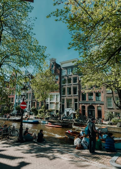 A narrow Amsterdam canal edged by brick townhouses and leafy trees, with moored boats on the water and people sitting along the stone quays on both sides under a clear sky