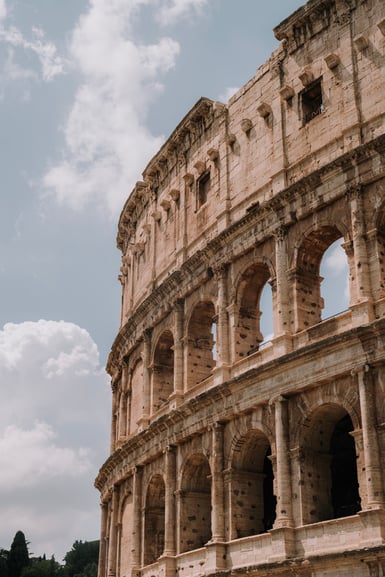 Low-angle view of the Colosseum's curving travertine facade in Rome, three tiers of arches and a solid upper storey pocked with holes, against a partly cloudy sky