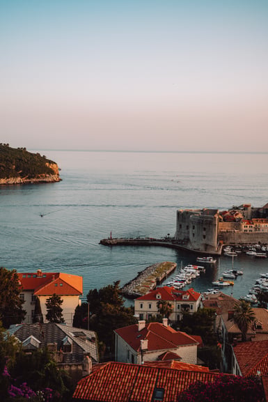 Elevated view over a small harbour on the Adriatic at dusk, with a stone fortress and breakwater enclosing moored boats, terracotta rooftops in the foreground and a pale sky above flat sea