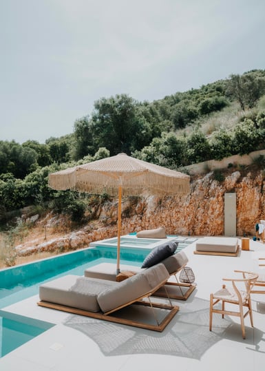 A small swimming pool with two wooden sun loungers and a fringed cream parasol on a pale terrace, backed by an ochre rock cut and green hillside scrub