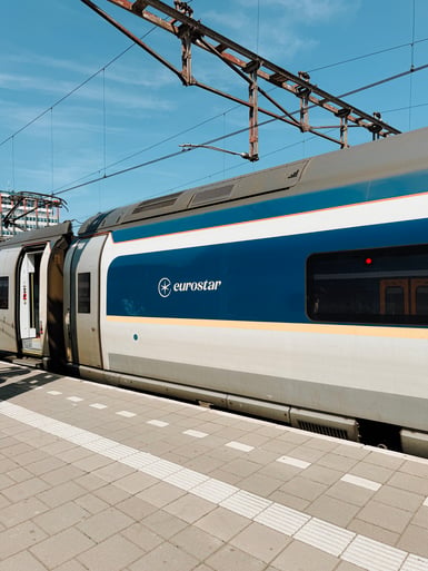 A blue and grey Eurostar train with an open door stands at an empty tiled platform under overhead catenary wires and a clear blue sky