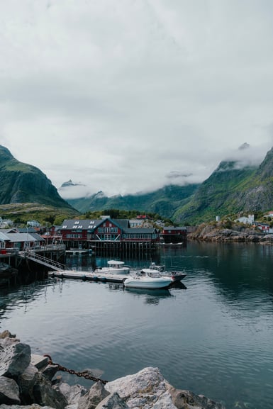 A small harbour in Lofoten with red timber buildings on stilts, white motorboats moored at a floating dock, and green mountains half-hidden by low cloud