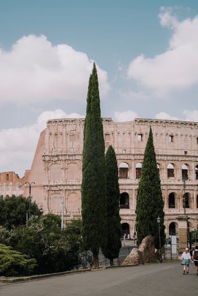 The Colosseum in Rome seen from a nearby road, its tiers of arches partly hidden behind three tall cypress trees, with oleander bushes, street lamps and a couple walking on the asphalt