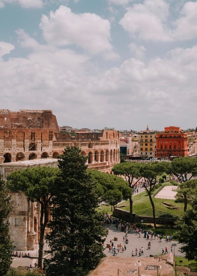 Elevated view of the Colosseum in Rome under a cloudy blue sky, with tall pines in the foreground, lawns, ochre buildings and small groups of visitors below