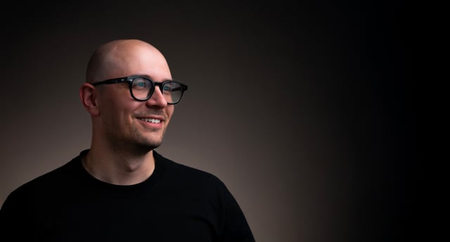 Studio portrait of a bald man with a short beard, thick black-framed glasses and a black t-shirt, smiling and looking off to the left against a dark brown background