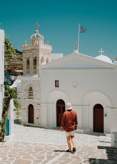 A man in a rust-coloured shirt, shorts and pale cap walks along a paved lane toward a whitewashed church with arched doors, a tiered bell tower and a Greek flag against clear blue sky