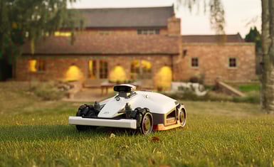 Mammotion Luba 2X robotic lawn mower on freshly cut grass in front of a warm brick countryside home at sunset.