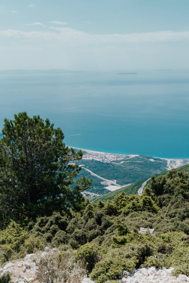 View from a scrub-covered limestone hillside with a pine tree at left, down over a coastal village and pale beach to a wide blue sea with hazy islands on the horizon