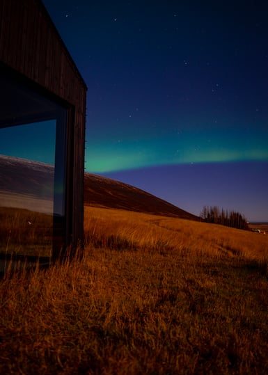 A dark timber cabin with a large glass window stands at left on a slope of dry golden grass, with a low hill, a line of trees and a green aurora band under a star-filled blue sky