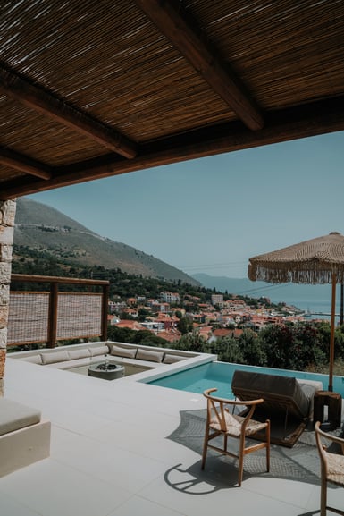 Shaded terrace with a reed-and-timber roof, a small pool, low white sofas and a fringed parasol, looking out over a red-roofed coastal village and hills