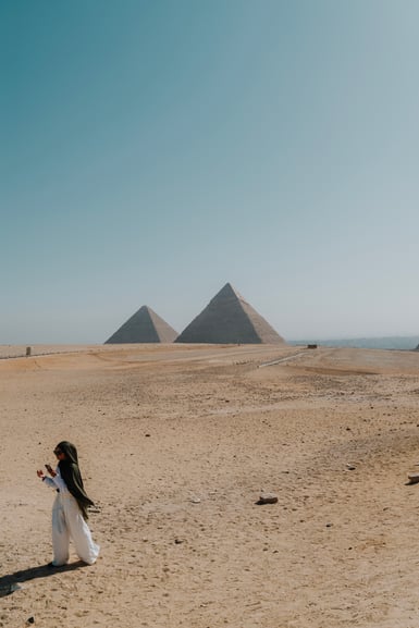 A person standing in a sandy desert with two pyramids in the distance under a clear sky.