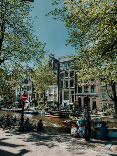 A tree-shaded Amsterdam canal with small moored boats, brick gabled houses along the far bank, and people sitting on the quayside beneath a no-entry sign