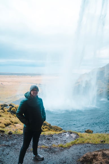 A man in a dark puffer jacket, grey beanie and glasses stands on a wet gravel path beside a tall waterfall, with flat farmland visible beyond the spray