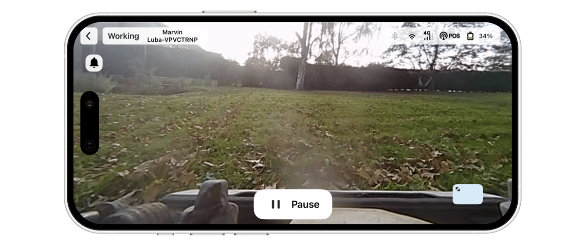A smartphone held horizontally shows a robot mower's live camera view of a leaf-strewn lawn with trees beyond, overlaid with a "Working" status label and a "Pause" button