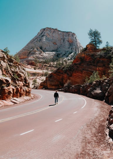 A person stands alone on the centre line of an empty curving road cut through red sandstone, with a pale domed sandstone peak rising ahead under a clear sky
