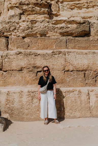 A woman wearing sunglasses stands against a large stone wall at an ancient site.