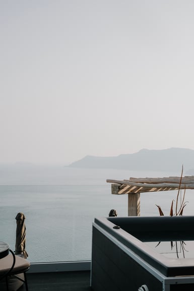 A terrace with a dark hot tub, striped wooden pergola posts and a wicker chair overlooking a pale grey sea, with a faint headland on the hazy horizon