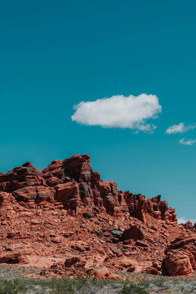 A low ridge of eroded red sandstone with scattered green desert shrubs at its base, beneath a deep blue sky holding a single small white cloud