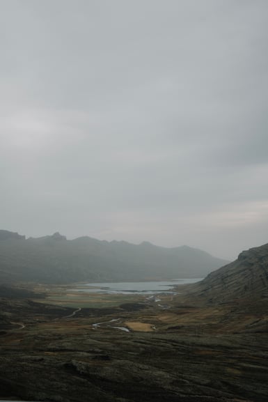 A wide overcast valley with a pale shallow lake, meandering river, small green fields and a gravel track, ringed by low ridges fading into mist under a grey sky