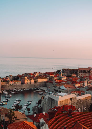 Elevated view over a walled coastal old town with terracotta rooftops and a small harbour of moored boats, the flat sea and pale pink sky filling the upper half