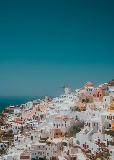 White and pastel cubic houses with domed churches and a windmill stacked down a steep hillside above the sea, beneath a large expanse of clear blue sky
