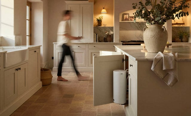 A cream-toned kitchen with stone floor tiles, a blurred person walking past cabinetry, and an open island cupboard revealing a white cylindrical water softener unit