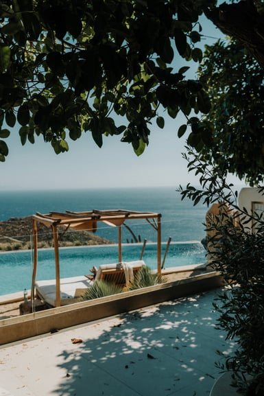 Dark leaves frame the top of the frame above a pale stone terrace, a wooden pergola shading a daybed beside an infinity pool, with dry hillside and open sea behind