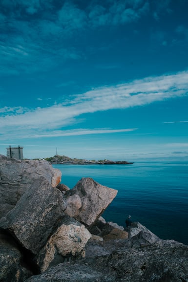 Large pale boulders in the foreground of a rocky coastline, with a lone figure sitting among them beside calm blue sea, a low headland and mast on the horizon under a streaked sky
