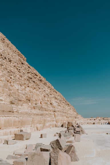 A large stone pyramid ruin with scattered blocks in the foreground beneath a clear blue sky.