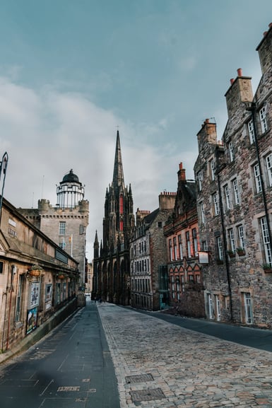 A cobbled street in Edinburgh's Old Town lined with tall stone tenements, leading toward a dark Gothic spire and a domed tower under a pale cloudy sky