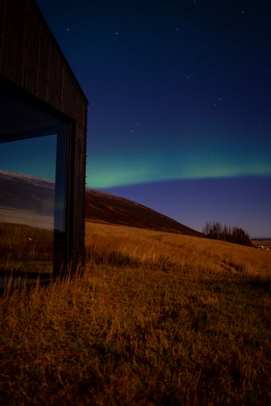 A dark timber-clad cabin with a large glass window stands at the left edge of a grassy hillside at night, with a faint green aurora band and stars over a bare hill and a line of conifers