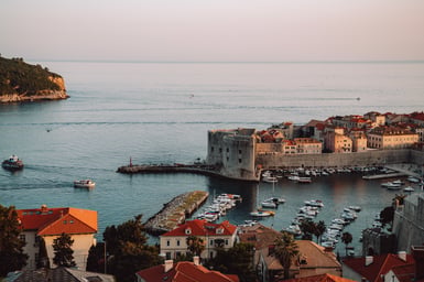 Elevated view over Dubrovnik's old harbour at dusk, with a stone fortress tower, breakwater, moored white boats, terracotta rooftops in the foreground and a flat pale sea beyond