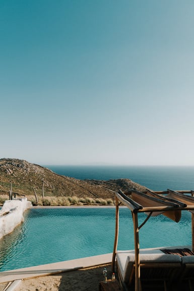 An infinity pool with a white rendered edge sits above a dry scrub hillside and the sea, with a timber-framed canopy, loungers and a small table with bottles at right