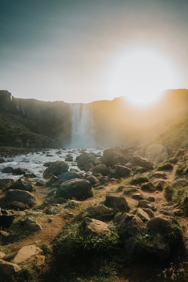 A waterfall drops from a cliff into a boulder-strewn river, with low sun burning over the right-hand ridge and a dirt path crossing grassy rocks in the foreground