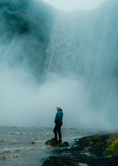 A person in a blue jacket and knit hat stands on wet dark rocks at the foot of a wide waterfall, looking up through heavy mist