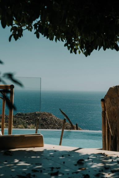 An infinity pool edge seen past a wooden pergola post and glass balustrade, with a rocky headland and the Aegean beyond, framed by dark overhanging leaves