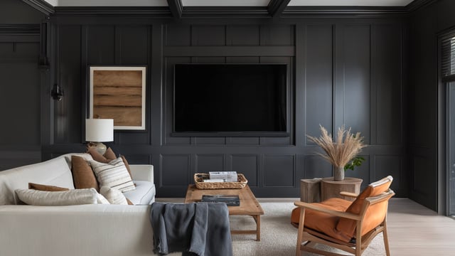 A living room with charcoal-grey panelled walls and a large flat-screen television set into a wood-framed recess, facing a pale sofa with cushions, wooden coffee table and leather armchair
