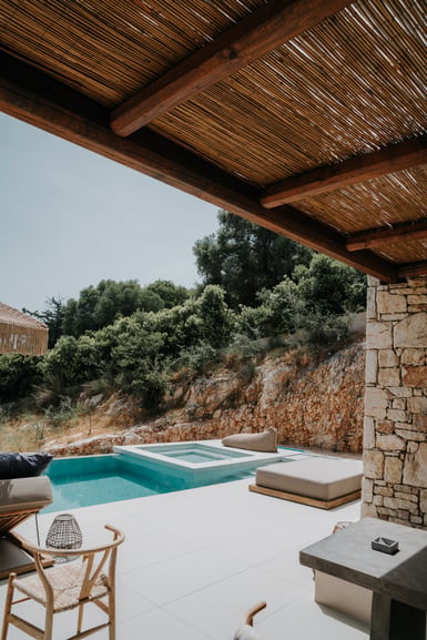 A reed-covered pergola on timber beams shades a pale stone terrace with a small pool, cushioned daybeds, a woven chair and a dry-stone wall, with green hillside behind